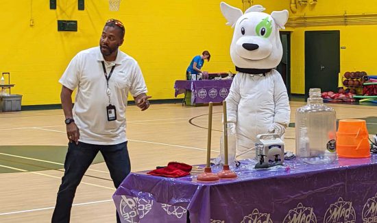 Scientist from Mad Science VA next to Sparky the Noblis Dog and a table full of science equipment during a demo for kids at STEM camp.