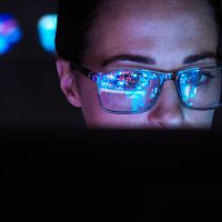 a woman's face shows partially from behind a computer screen, an image of code is reflected in her glasses