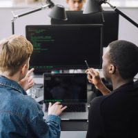 Two colleagues sit at a desk in front of a set of computer screens discussing AI algorithm development.