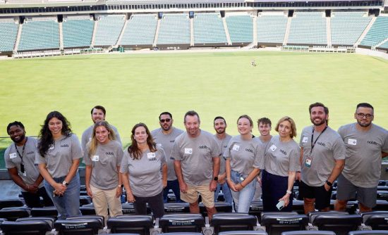 A group of 13 Noblis employees stand in a row between the seats of the Philadelphia baseball team stadium with the field in full view behind them.