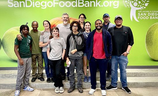 A group of Noblis employees and their families stand together in front of a wall with a Feeding San Diego food bank banner.