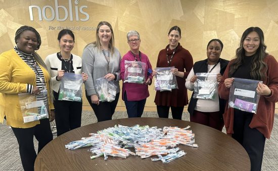 Seven Noblis employees standing around a table with supplies for hygiene kits. Each person holds an assembled kit up for the camera.