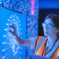 A woman in glasses looking and pointing at a large screen showing site surveillance graphs and data.