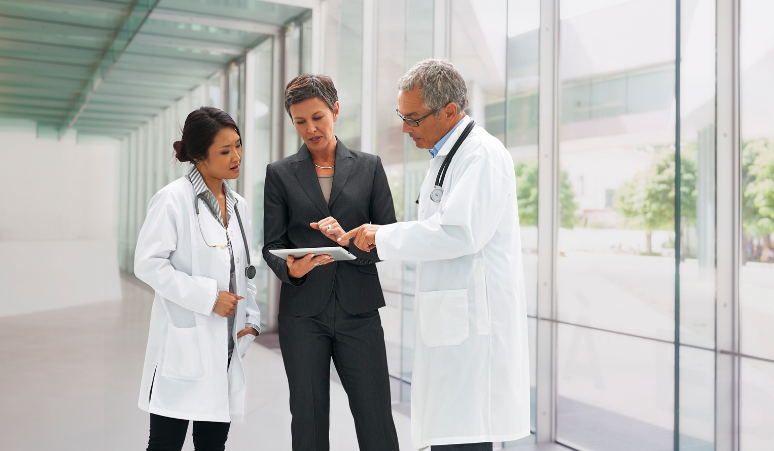 two doctors and a program leader stand in a hospital hallway looking at information on a tablet