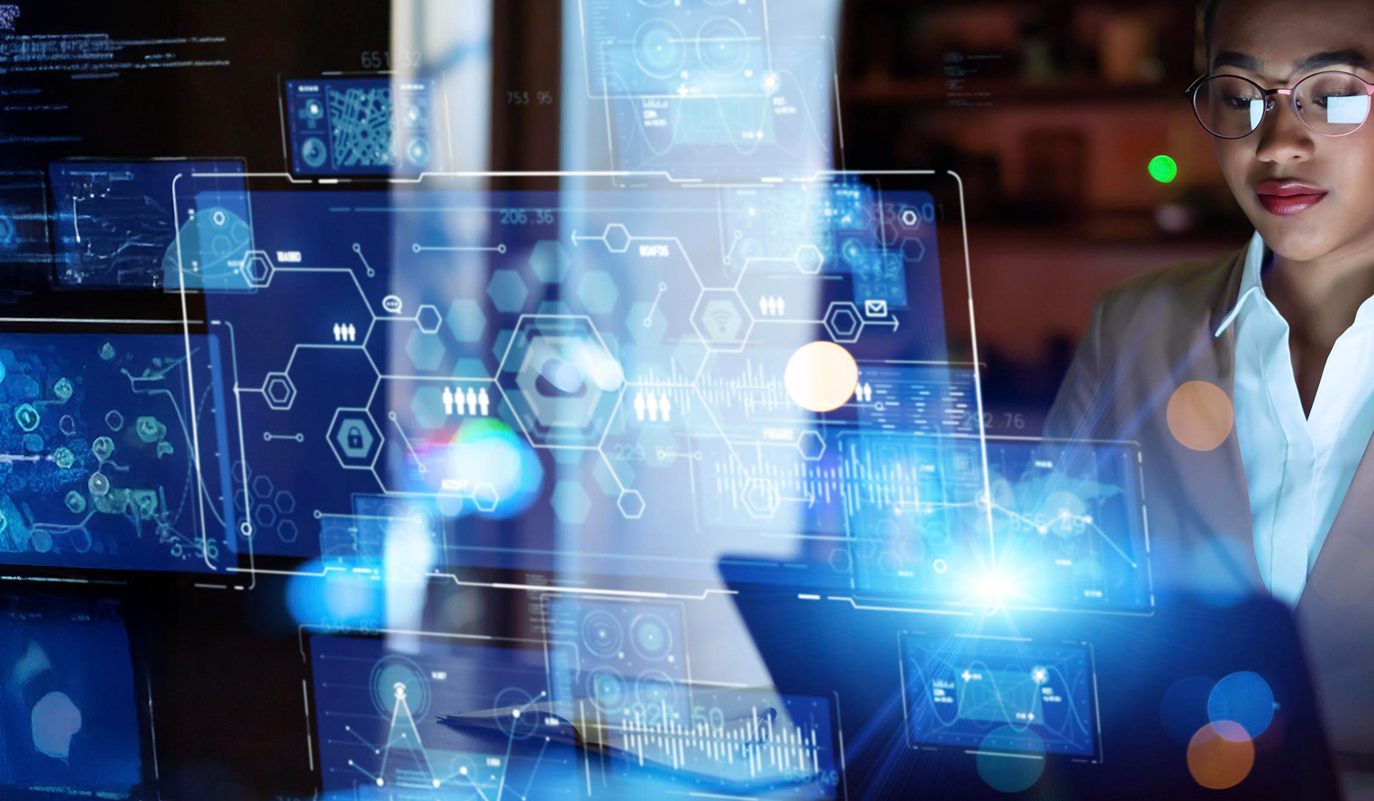 a professional looking woman in glasses looks down at a laptop, to the front of her glassy, digital images of data dashboards float in the air