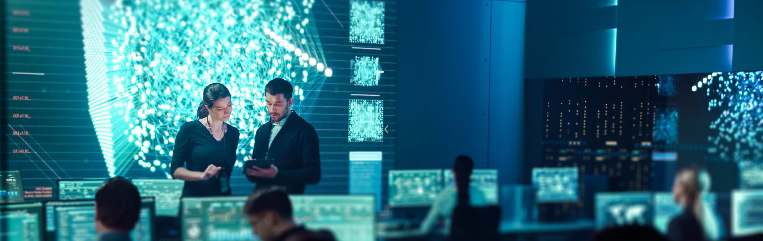 two homeland security professionals talk in a control room with many screens on the wall and at stations where other analysts work