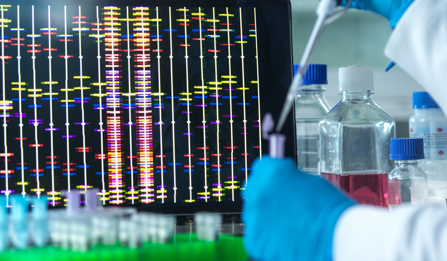 a screen in a lab shows genomic data and a lab worker wearing gloves is putting liquid in a small vial