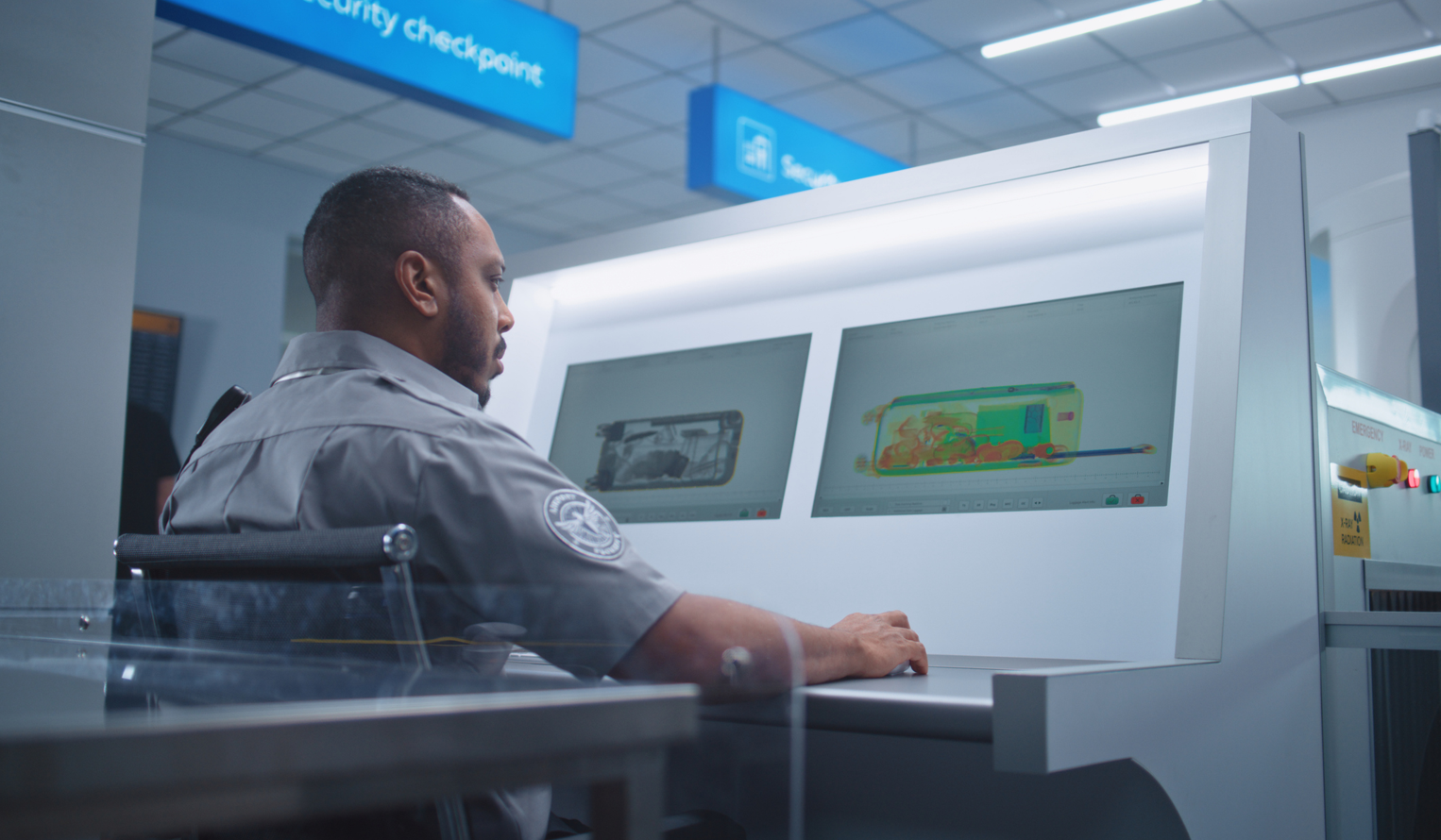 A TSA agent sits at a monitor reviewing security checkpoint scans of luggage
