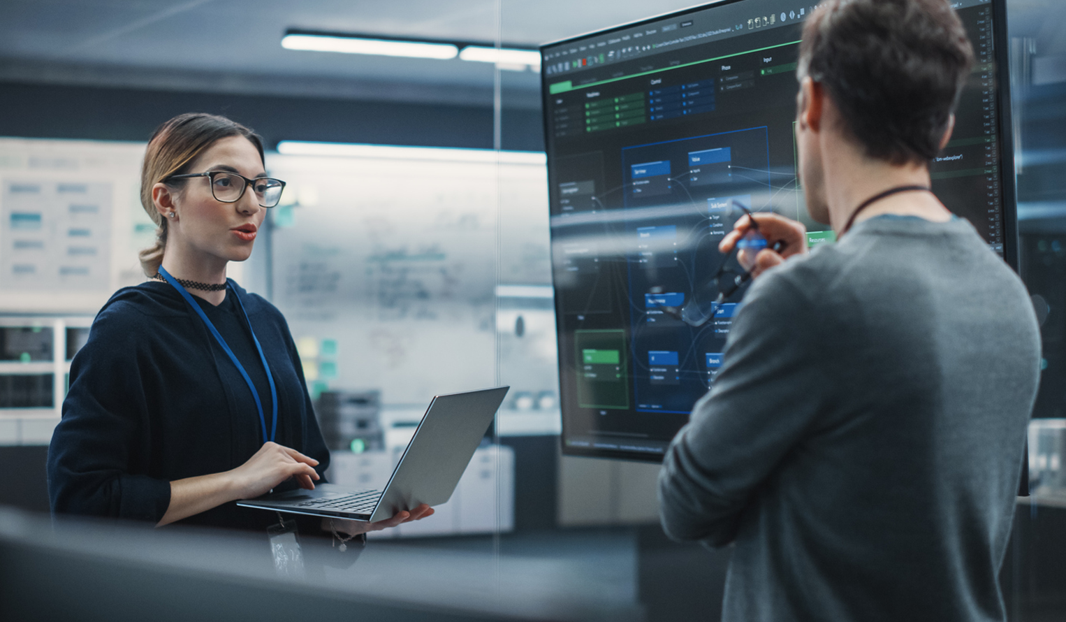 two systems engineers review and discuss a digital engineering plan on a big screen
