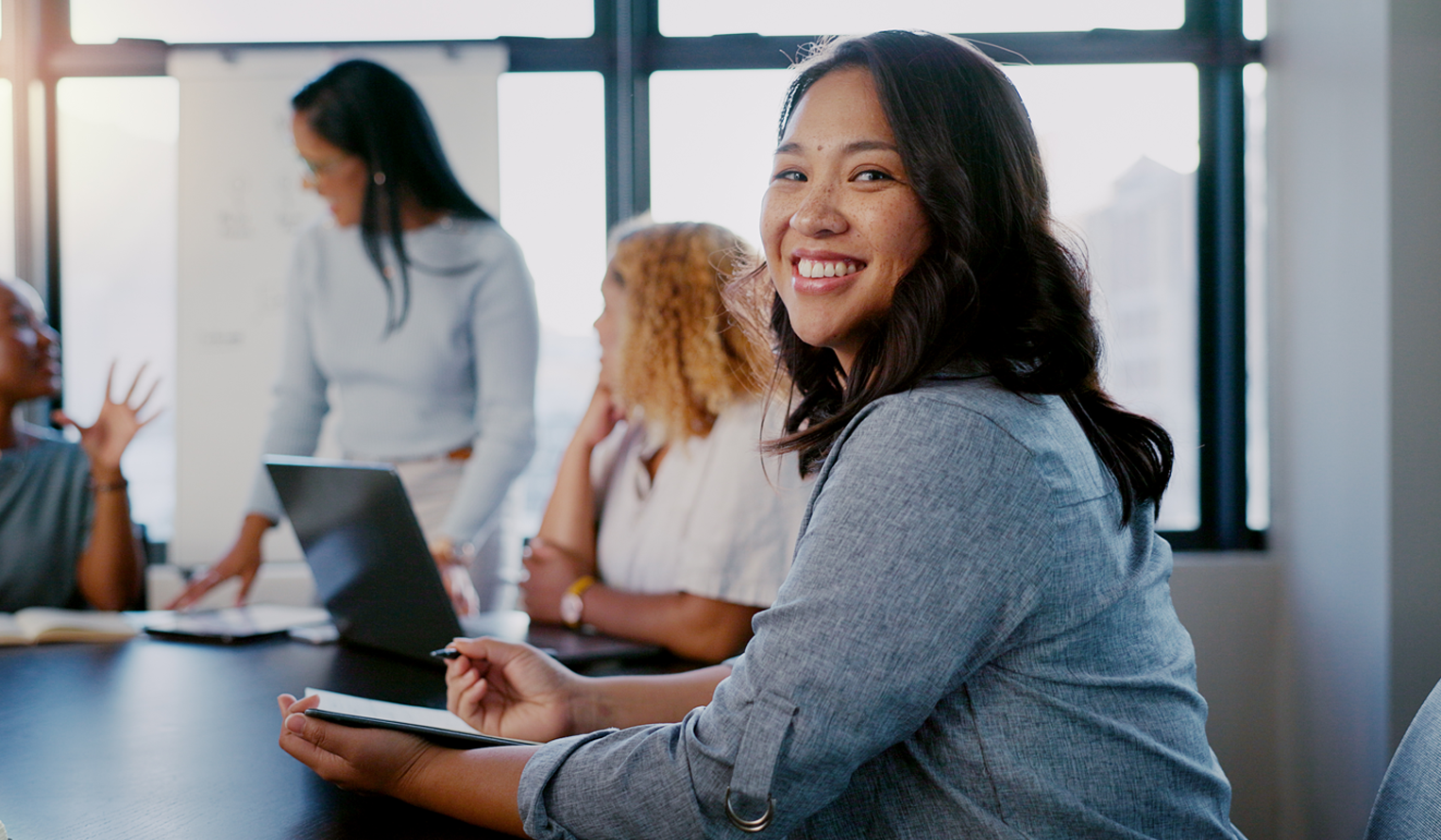 a group of employees sits talking in an office conference room. the woman at the foreground smiles at the camera.