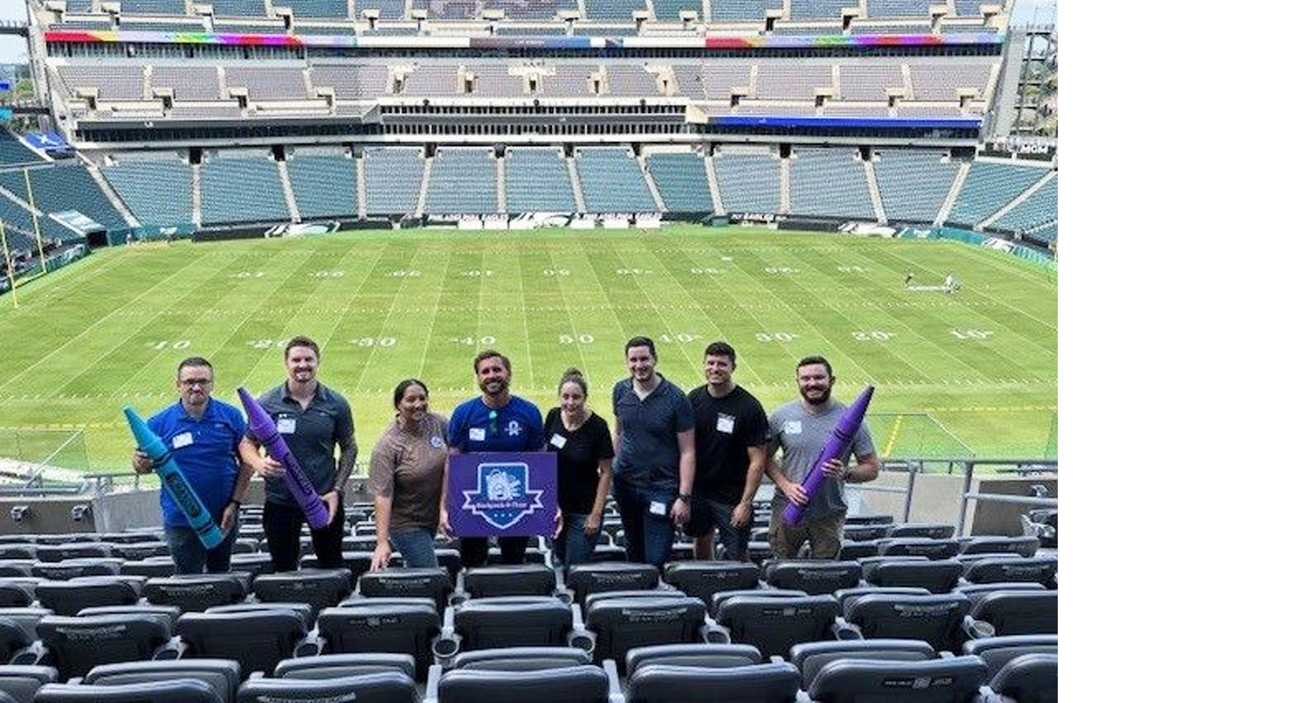 Noblis MSD team volunteers together in the stands at Lincoln Park stadium holding backpacks and inflatable crayons.