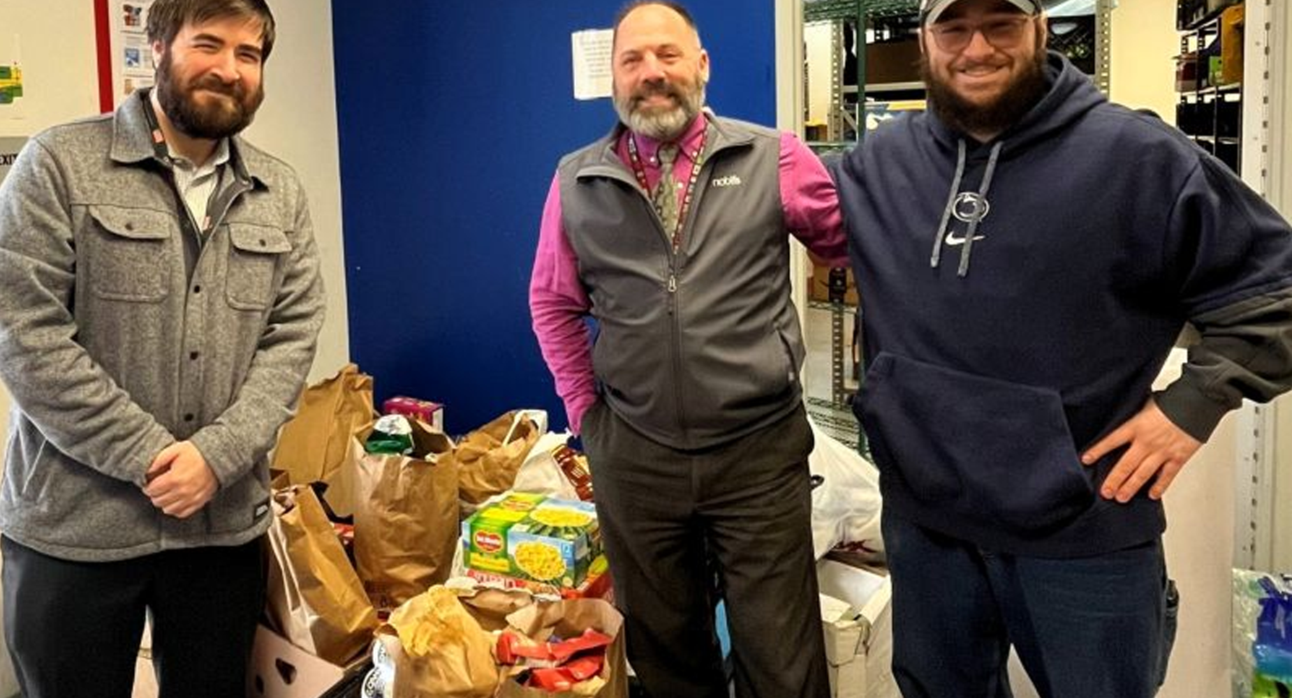 Two Noblis employees stand with the LCAC director in front of food collected for people in need.